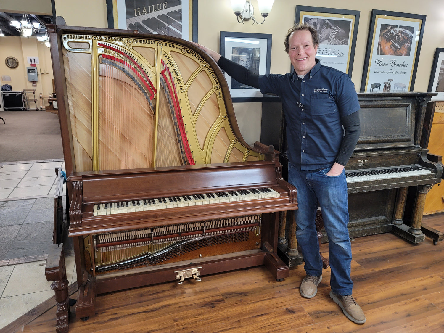 Brigham Larson smiling while standing between an unusual, verti-grand Schimmel & Nelson upright piano open-frame vintage upright piano with exposed strings and an antique wood-grain piano inside the Brigham Larson Pianos family heirloom piano restoration shop in Orem, UT.