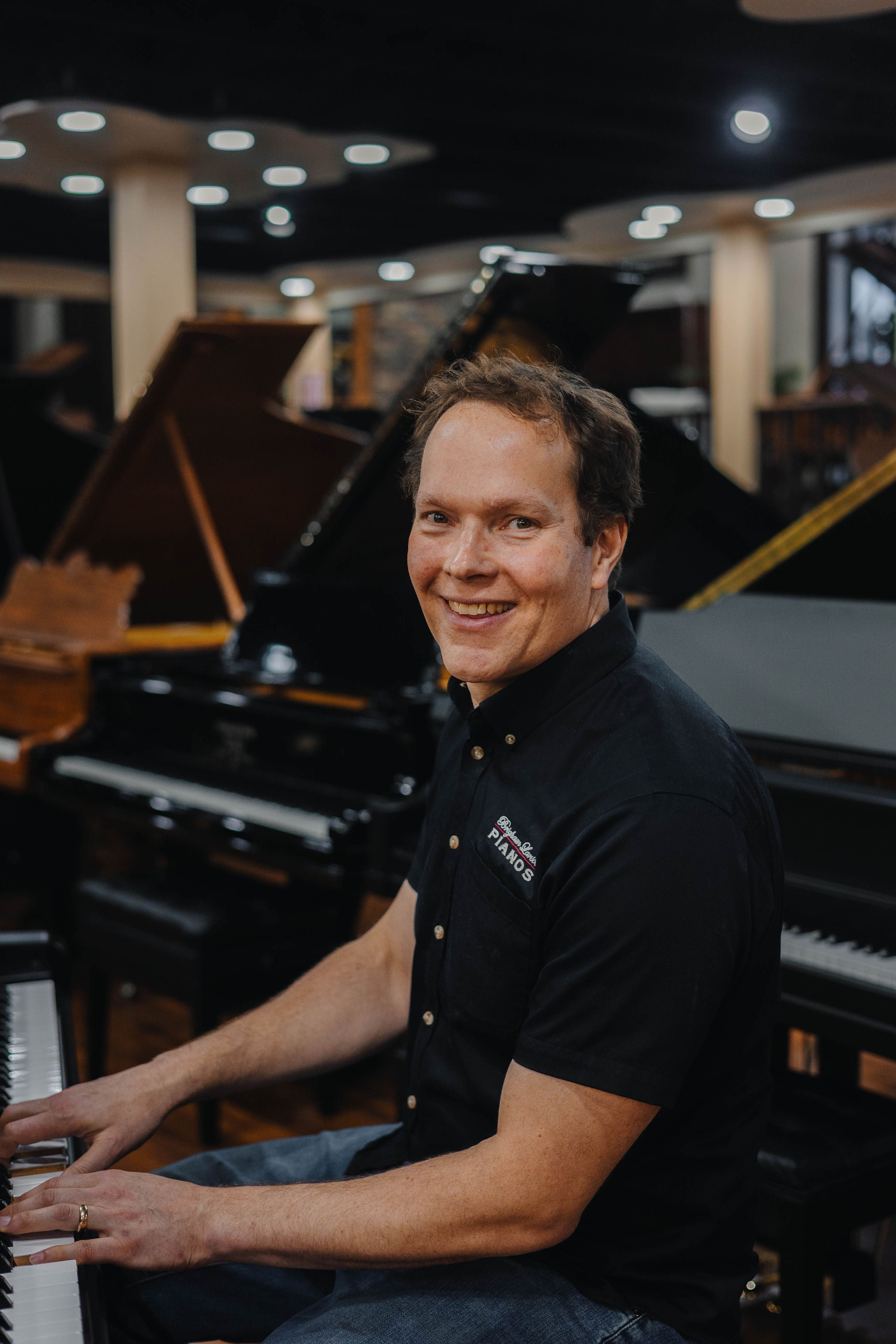 Brigham Larson, Registered Piano Technician, playing a piano in the Orem, Utah Brigham Larson Pianos piano store sales showroom with pianos in the background.