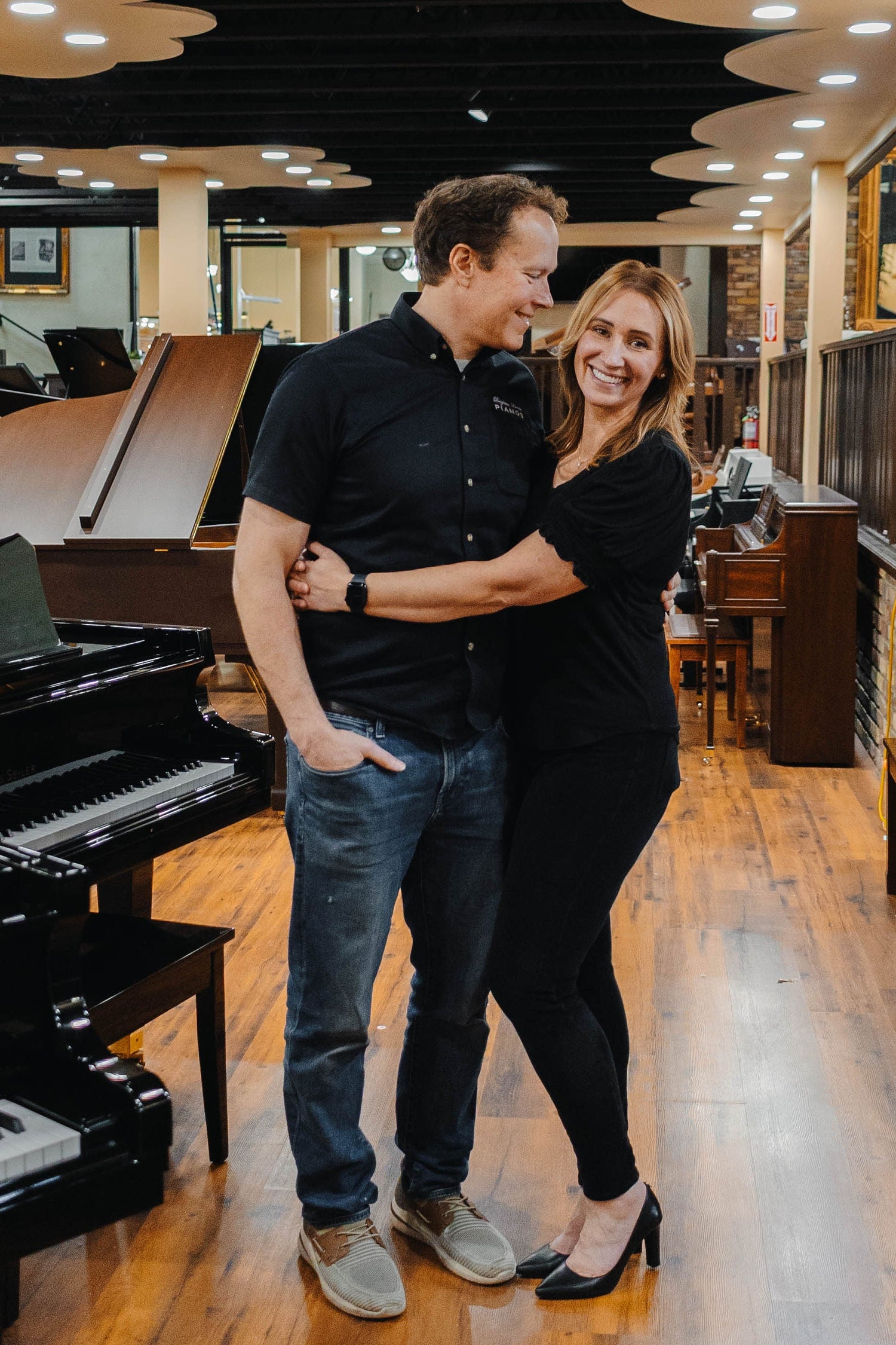 Brigham & Karmel Larson embracing in a piano store with grand and upright pianos, at Brigham Larson Pianos in Orem, UT.