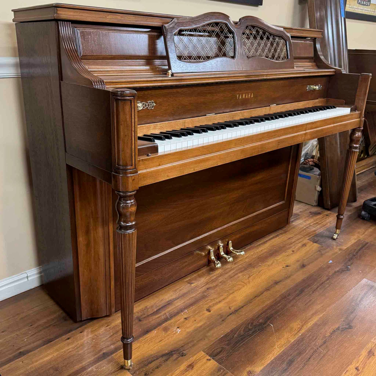 Wooden Yamaha M series console piano on a wooden floor with a beige wall in the background in the Brigham Larson Pianos piano restoration shop. 