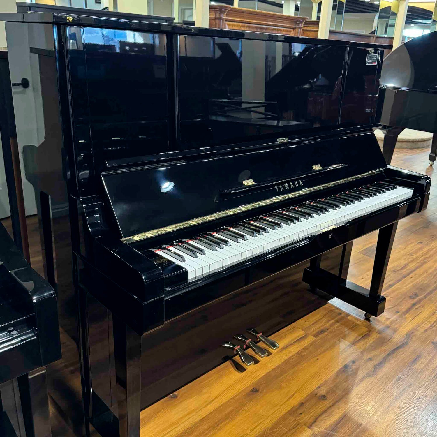 Polished Black Yamaha YUX upright piano on a wooden floor in a piano sales floor showroom setting