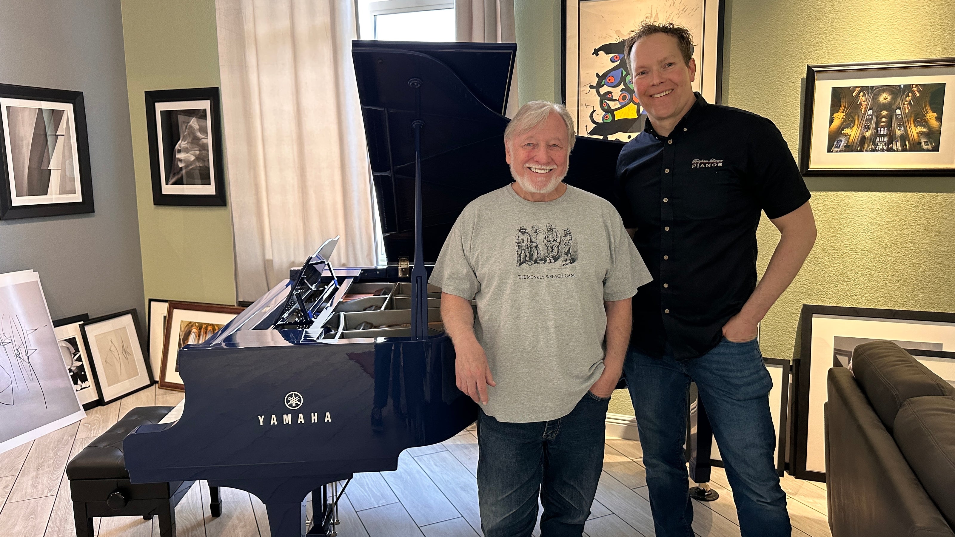 Brigham Larson standing next to a customer and a polished navy blue Yamaha grand piano with its lid propped open, delivering and setting up the fully restored heirloom piano the customer's home in Nevada. 