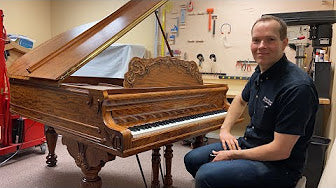 Brigham Larson sitting on a bench next to an antique wood-grain grand piano being worked on inside the Brigham Larson Pianos family heirloom piano restoration shop in Orem, UT.