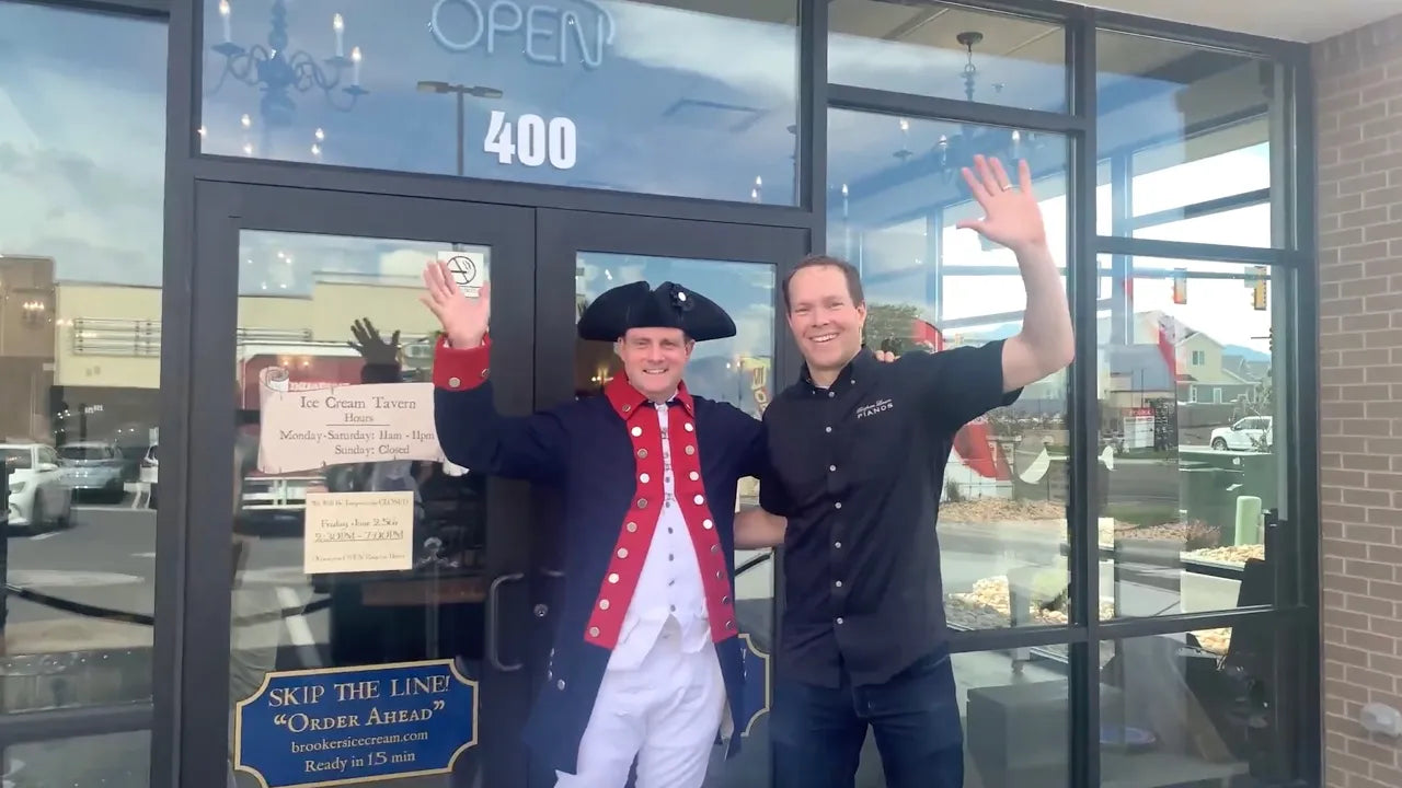 Brigham Larson waving alongside a man dressed in historical colonial attire outside the glass storefront entrance of Brookers IceCream with white Hailun Grand Piano in the background. 